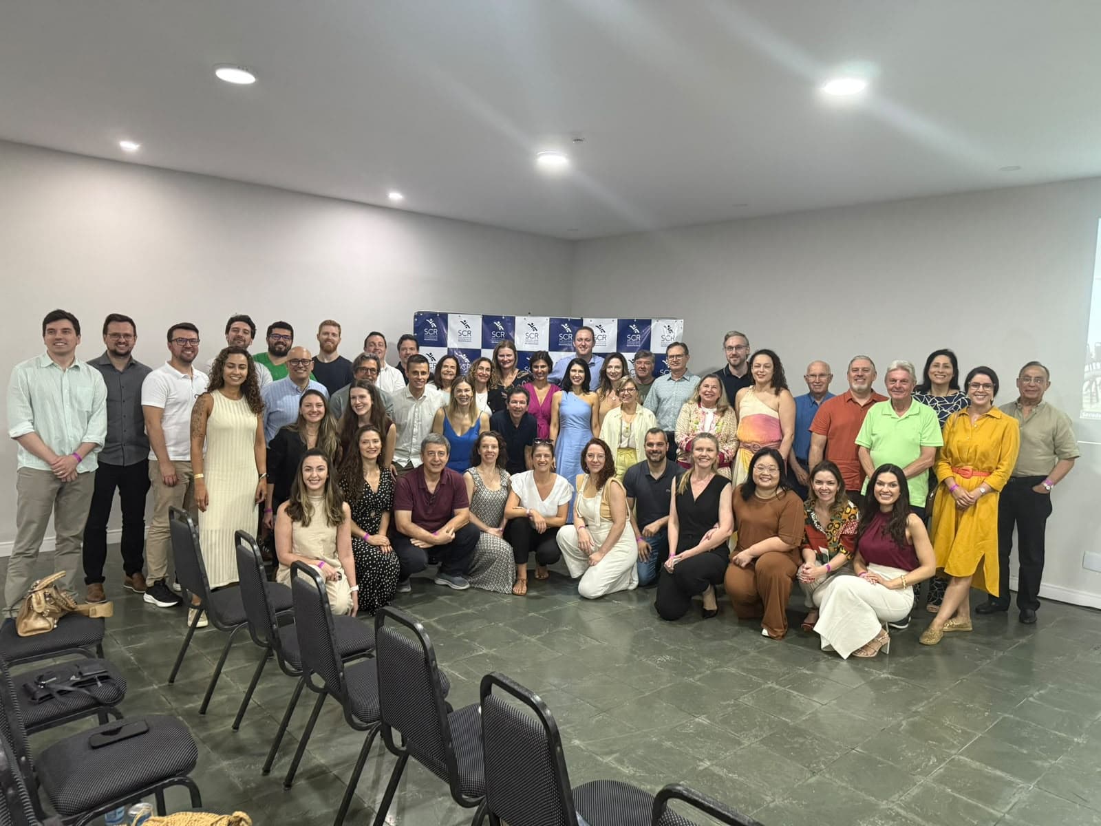 Grupo de associados da SCR reunidos em frente ao painel da sociedade durante um evento.